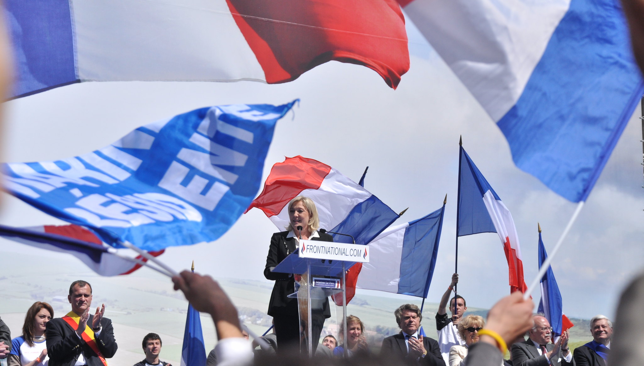 An image of Marine Le Pen at a podium in front of a rallying crowd.